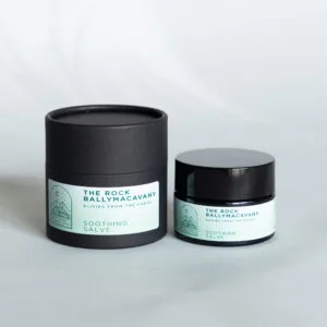 A black cylindrical container and a small black jar, both labeled Soothing Salve with a light blue mountain and crescent moon design, sit on a light grey surface.