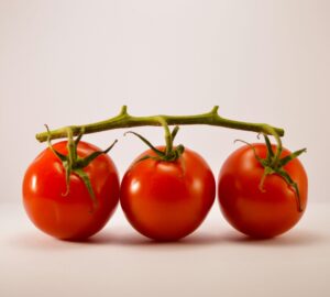 Three ripe Cherry Tomato – Gardeners delight are attached to a green vine and displayed against a plain, light background.