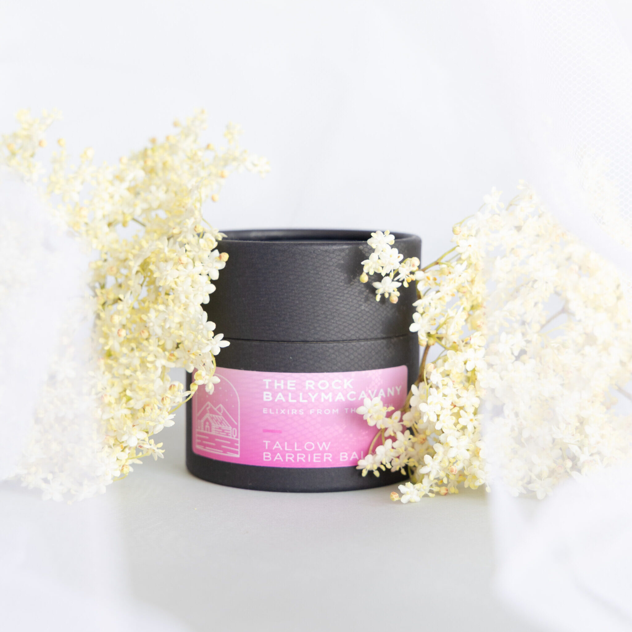 A black jar labeled Tallow Barrier Balm with a pink label sits beside a cluster of small white elderflowers on a soft white fabric background.