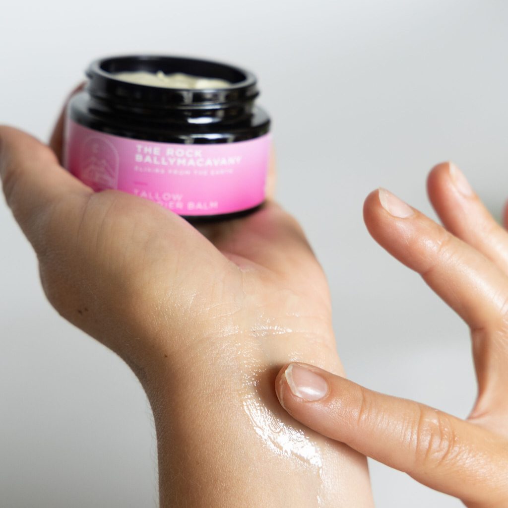 A person applies Tallow Barrier Balm from a pink-labeled jar onto their wrist with their finger, creating a glossy, moisturised finish on the skin.