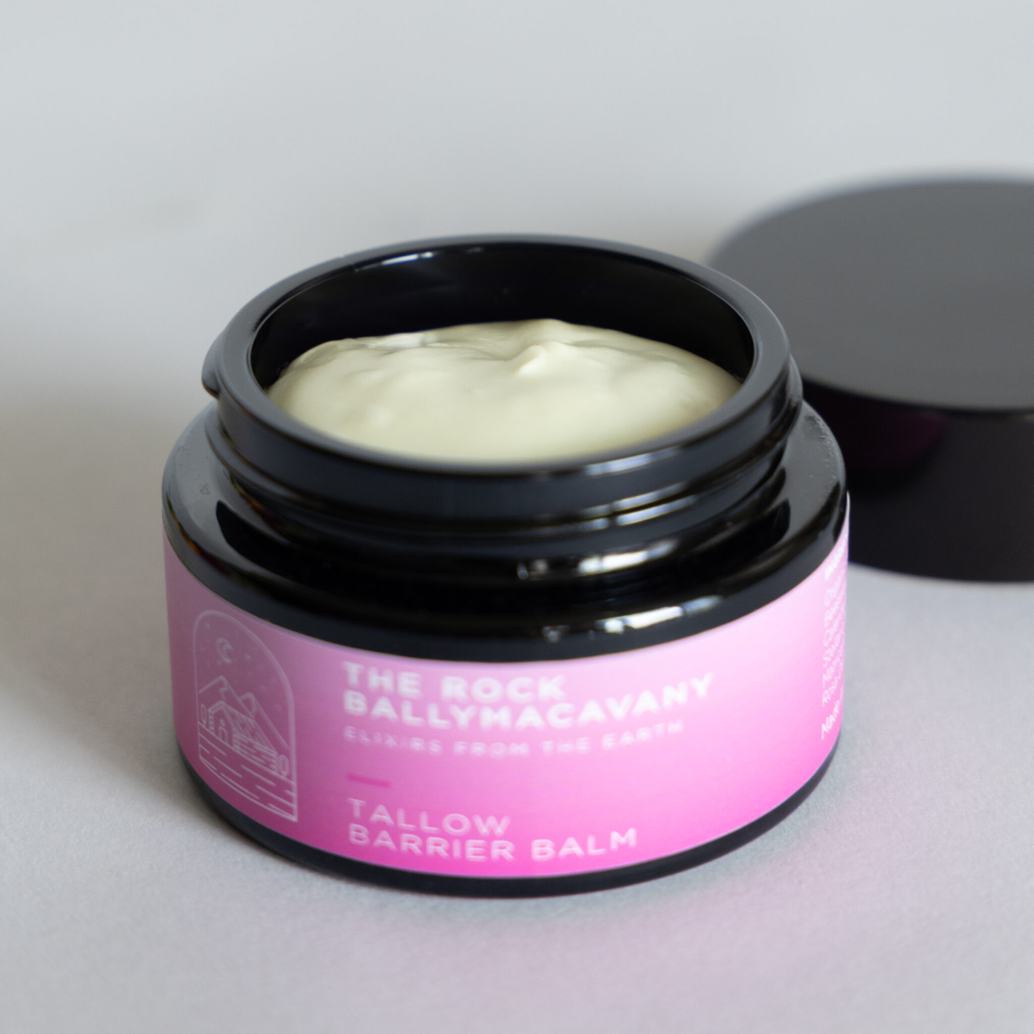 An open black jar of Tallow Balm with a pink label reveals creamy balm inside, while its black lid rests nearby on a light gray surface.