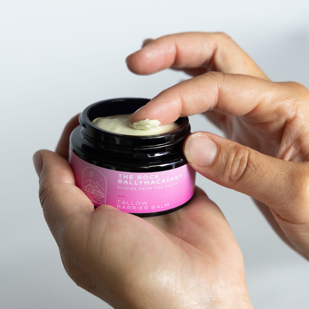 A hand holds an open jar of Tallow Barrier Balm as another hand scoops out some balm with a finger. The jar features a pink label with white text.