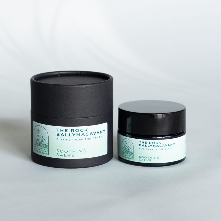 A black cylindrical container and a small black jar, both labeled Soothing Salve with a light blue mountain and crescent moon design, sit on a light grey surface.