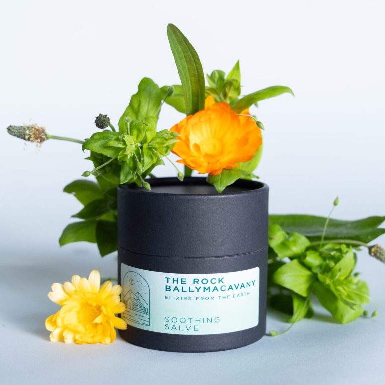 A black cylindrical container labeled Soothing Salve is surrounded by fresh green leaves and yellow flowers against a light background.