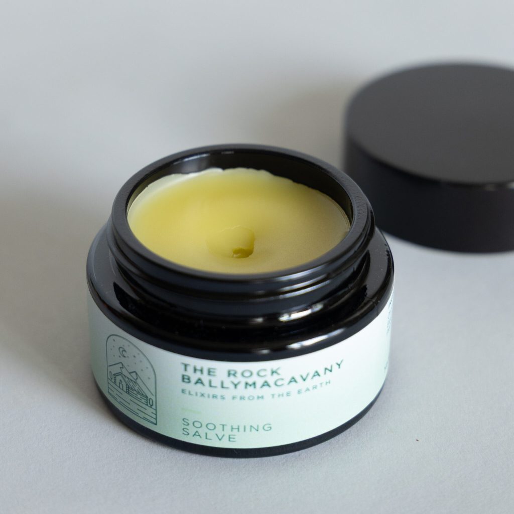 Open jar of Soothing Salve with yellow balm, labeled “The Rock Ballymacavany Elixirs from the Earth”; black lid beside it on a light gray background.