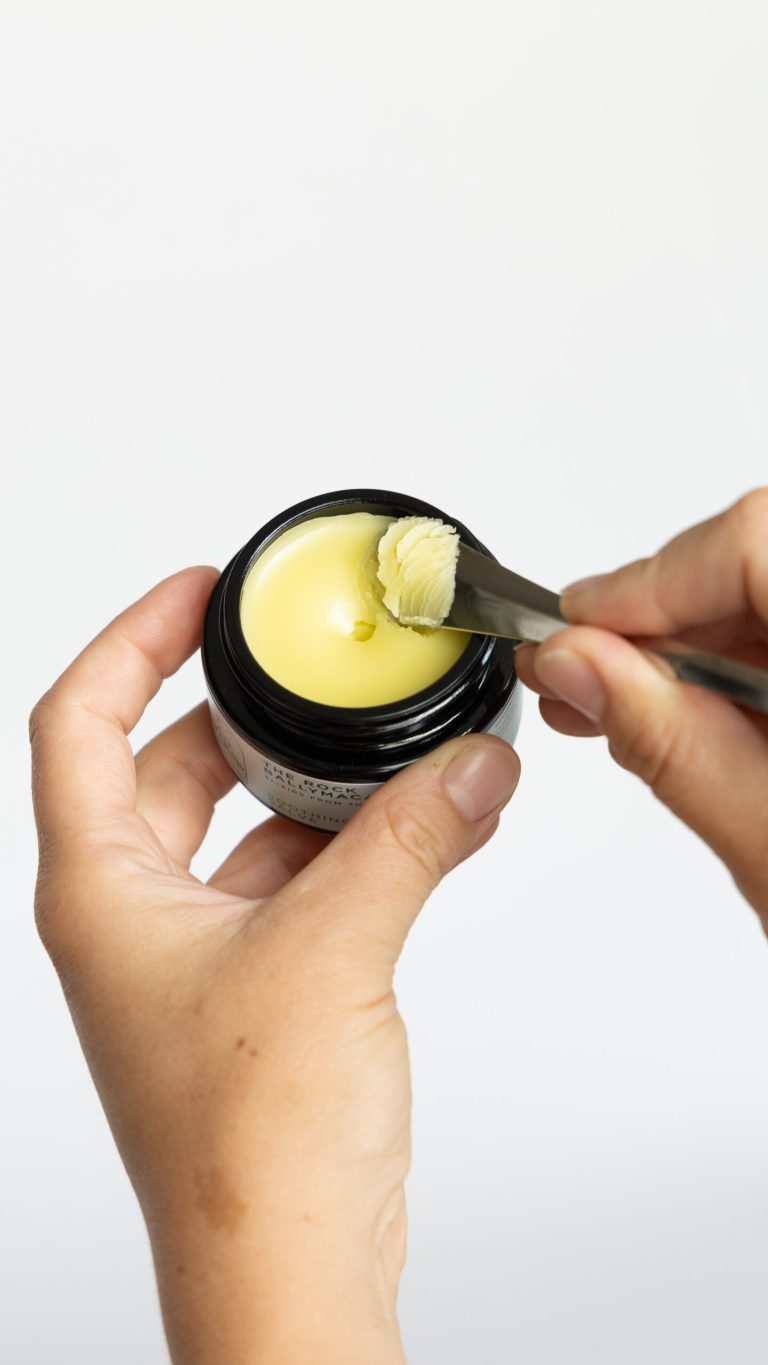 A person holds a small black jar of Soothing Salve, filled with creamy yellow herbal salve, and uses a metal spatula to scoop out a small amount against a plain white background.