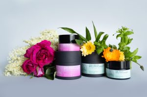 Two Tallow Balms and two Soothing Salves with pastel labels are arranged in front of fresh roses, calendula, and white blossoms, set against a light gray background.