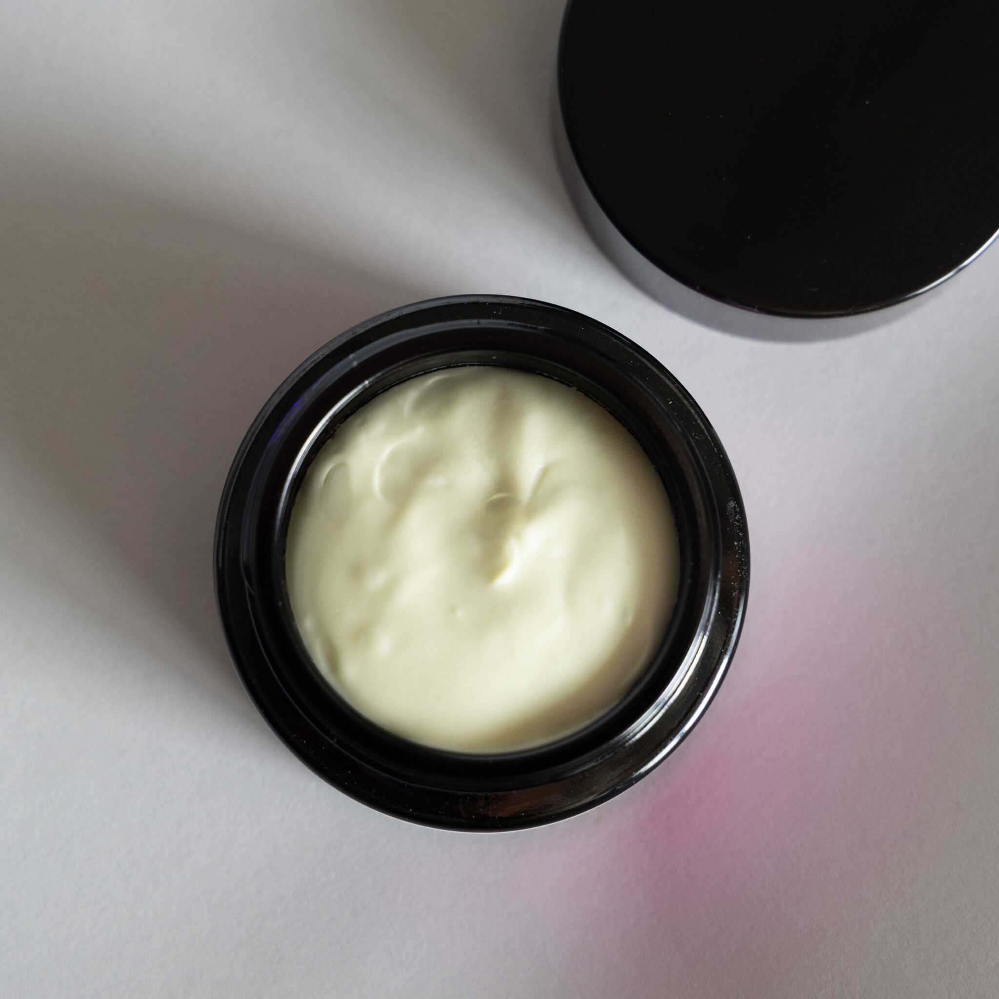 A black open container of Tallow Balm, filled with creamy light yellow balm, sits on a white surface with its lid placed nearby.