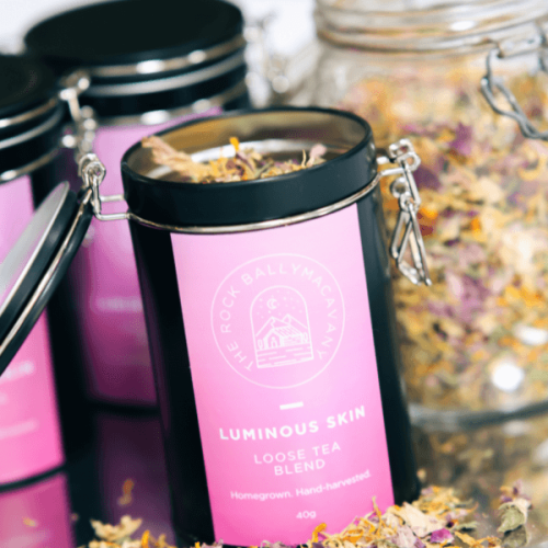 an open container of luminous skin tea in front of a clear glass jar of tea leaves and two more closed containers of luminous skin