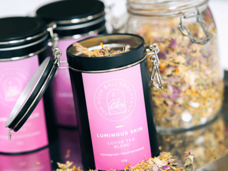 an open container of luminous skin tea in front of a clear glass jar of tea leaves and two more closed containers of luminous skin