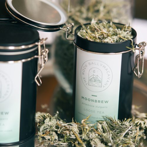 an open container of moonbrew herbal tea, with tealeaves in front of the container. the container is situated beside a closed container of moonbrew and in front of a glass jar of moonbrew tea leaves