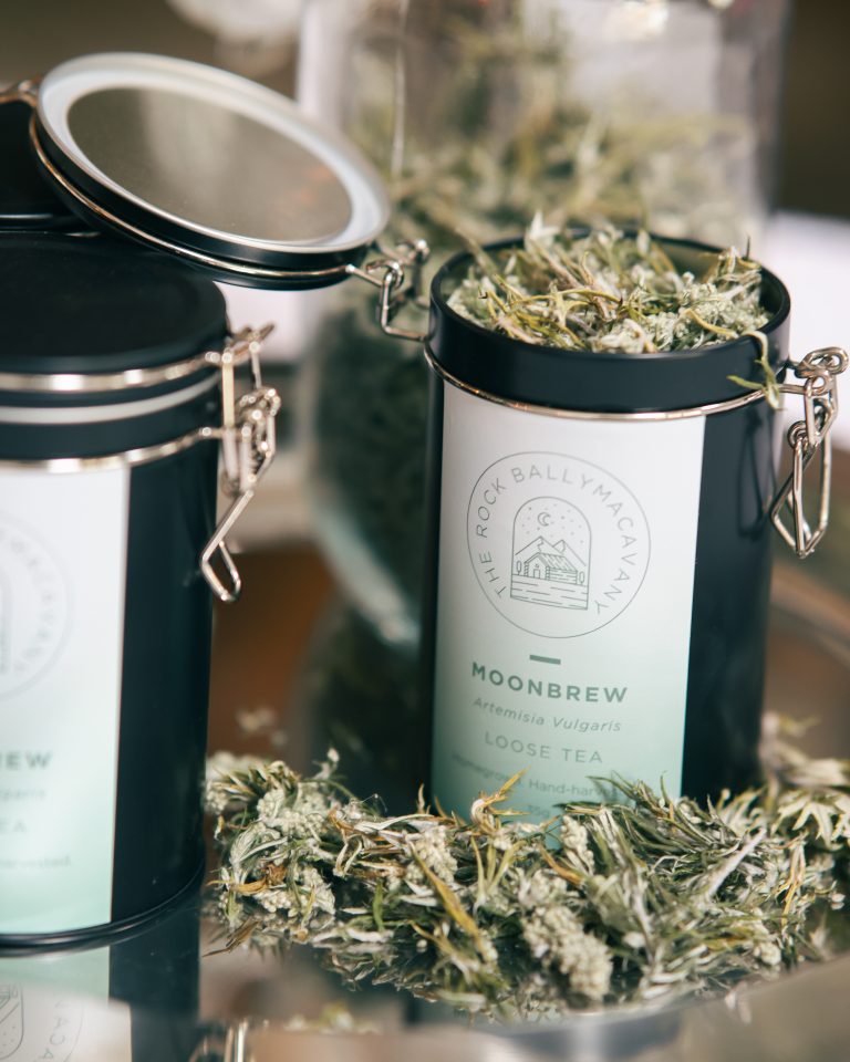 an open container of moonbrew herbal tea, with tealeaves in front of the container. the container is situated beside a closed container of moonbrew and in front of a glass jar of moonbrew tea leaves